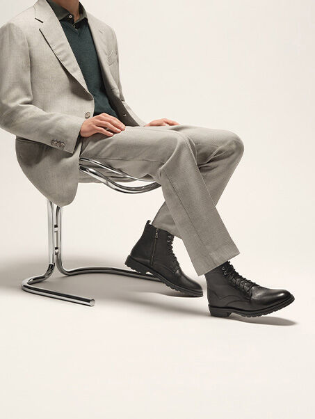 Man in a light grey suit with a shirt and V-neck jumper sitting on a metal armchair with lace-up ankle boots on his feet | GEOX | GEOX